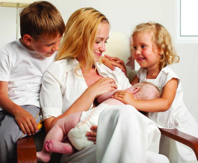 A mother breastfeeds while her two older children sit with her.